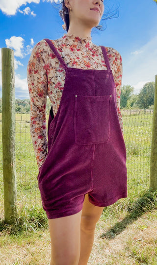 Plum corduroy overall shorts with front pocket styled over a floral mock-neck top in a sunny outdoor setting.