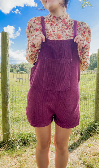 Plum corduroy overall shorts with front pocket, layered over a floral mock-neck top in a bright outdoor setting.