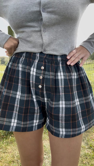 Woman wearing high-waisted black and blue plaid shorts with white and rust stripes and front buttons, cozy fall boutique style.