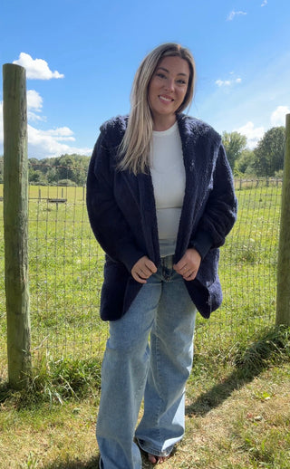 Woman wearing a navy blue teddy coat over a white top and light wash jeans in a sunny outdoor field.