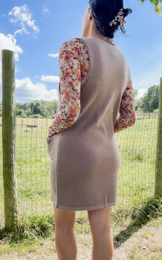 Back view of taupe sleeveless knit jumper dress layered over a floral long-sleeve mock-neck top in a sunny field.