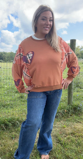 Burnt orange sweatshirt with plaid patchwork accents on the sleeves and front, styled with classic blue jeans for a cozy fall vibe.