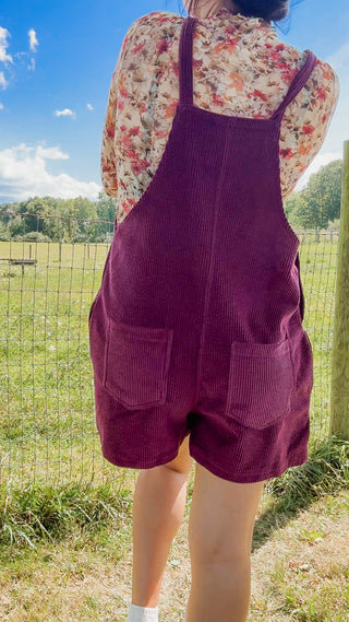 Back view of plum corduroy overall shorts with dual patch pockets, paired with a floral long-sleeve top.
