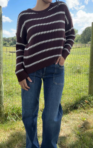 Brown knit sweater with white diagonal stripes paired with blue denim jeans in a sunny outdoor setting.