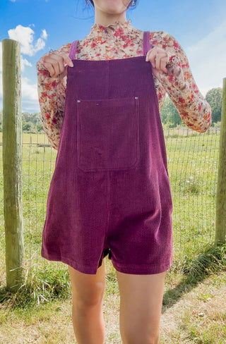 Plum corduroy overall romper with front pocket, styled over a floral mock-neck top in a sunny outdoor setting.