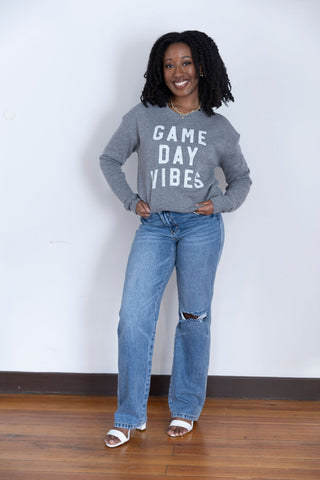 A smiling woman in a heather gray crewneck sweatshirt with bold white "Game Day Vibes" text, paired with classic blue jeans.