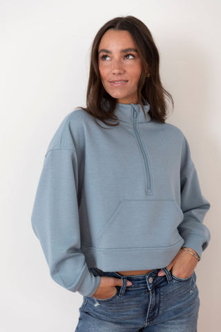 Woman wearing a cropped, slate blue quarter-zip sweatshirt with a kangaroo pocket, styled with distressed denim jeans