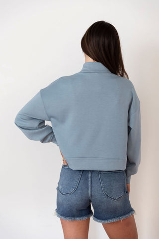 Woman wearing a cropped, slate blue quarter-zip sweatshirt with a kangaroo pocket, styled with distressed denim jeans