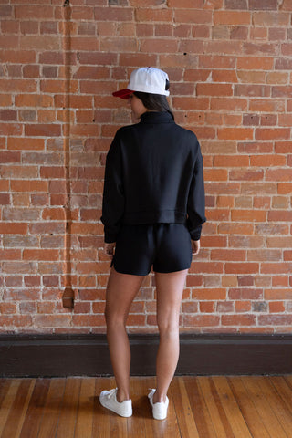 Woman wearing a black half-zip cropped sweatshirt with matching high-waisted shorts, styled with a trucker hat, against a brick wall