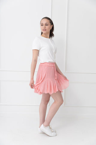 Model in white cropped top and pink pleated mini skirt with white sneakers, posing against minimalist white background