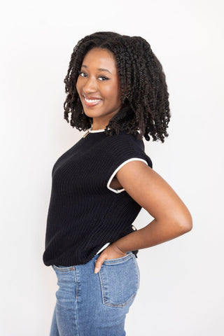 Woman wearing a black sleeveless knit sweater with contrast white trim and high-waisted blue jeans, posing against a neutral backdrop.