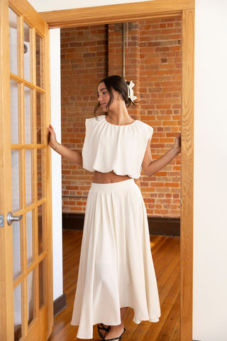 Woman wearing an elegant cream two-piece set with a flowy cropped top and high-waisted maxi skirt, standing in a doorway with brick wall backdrop.