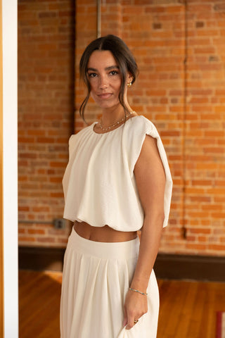 Woman wearing an elegant cream two-piece set with a flowy cropped top and high-waisted maxi skirt, standing in a doorway with brick wall backdrop.