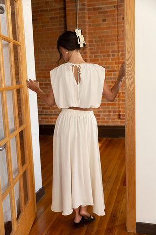 Back view of a woman in a cream two-piece set, featuring a tie-back cropped top and flowy maxi skirt, styled with a chic hair bow.