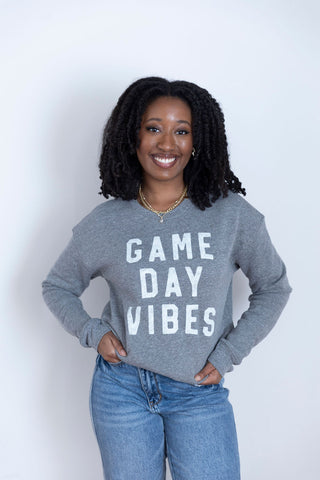 A smiling woman in a heather gray crewneck sweatshirt with bold white "Game Day Vibes" text, paired with classic blue jeans.