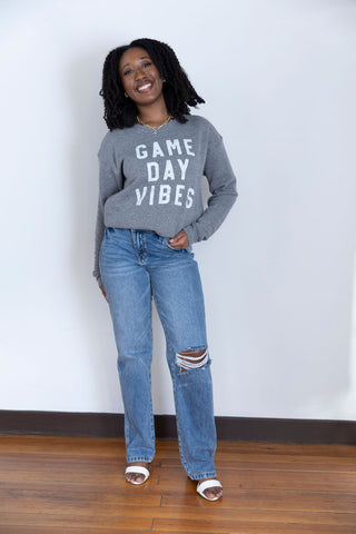 A smiling woman in a heather gray crewneck sweatshirt with bold white "Game Day Vibes" text, paired with classic blue jeans.