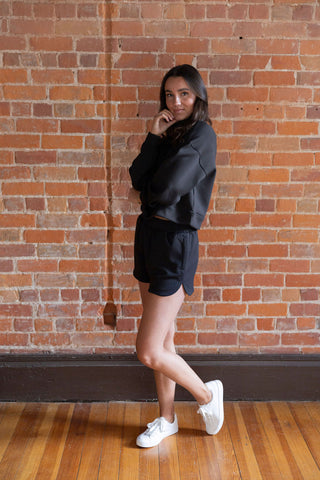 Woman wearing a black half-zip cropped sweatshirt with matching high-waisted shorts, styled with a trucker hat, against a brick wall