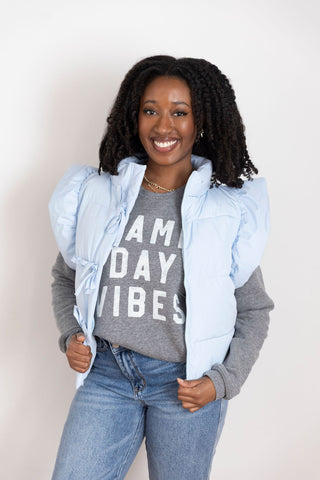 Model wearing a light blue puffer vest with ruffled shoulder details, styled over a gray graphic sweatshirt and high-waisted jeans.