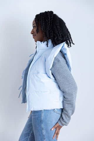 Model wearing a light blue puffer vest with ruffled shoulder details, styled over a gray graphic sweatshirt and high-waisted jeans.