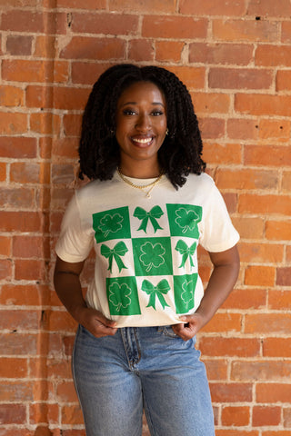 Women's cream graphic tee with green shamrock and bow print. Festive St. Patrick's Day t-shirt styled with casual denim jeans.