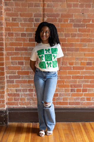 Women's cream graphic tee with green shamrock and bow print. Festive St. Patrick's Day t-shirt styled with casual denim jeans.